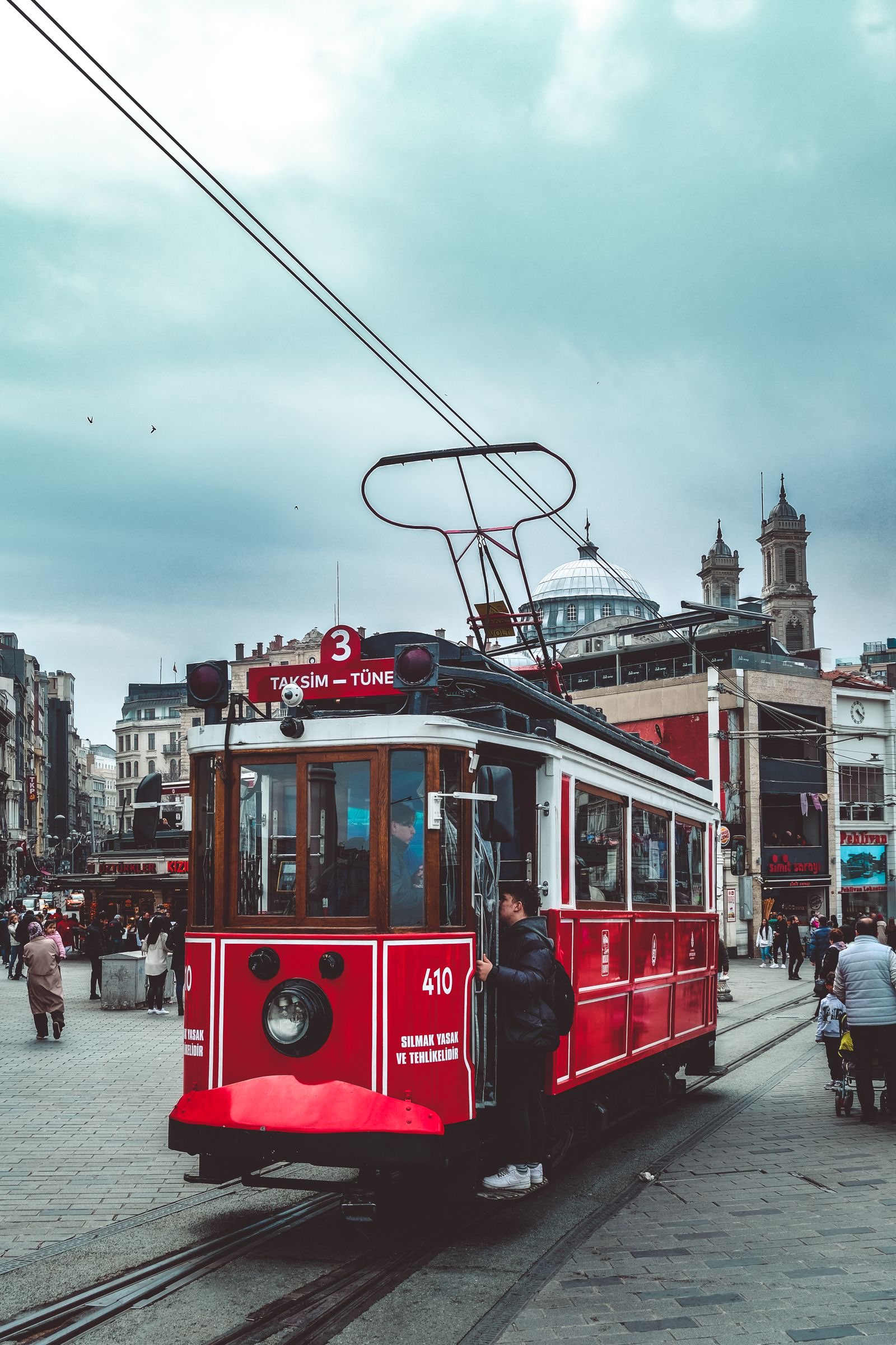 Red nostalgic tram marked Taksim-Tünel on cobbles at Taksim Square with pedestrians and a domed church behind, Istanbul