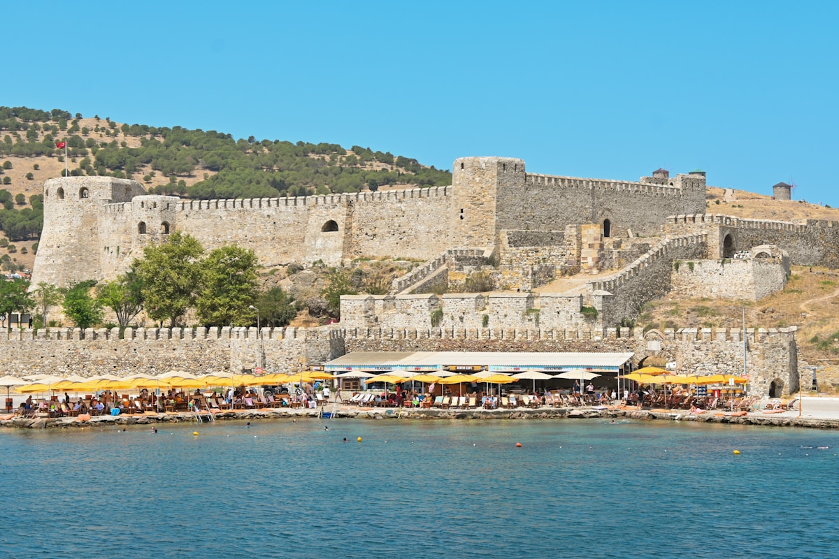 Stone fortress walls rising above a sandy beach on the Aegean coast of Turkey, with yellow beach umbrellas below